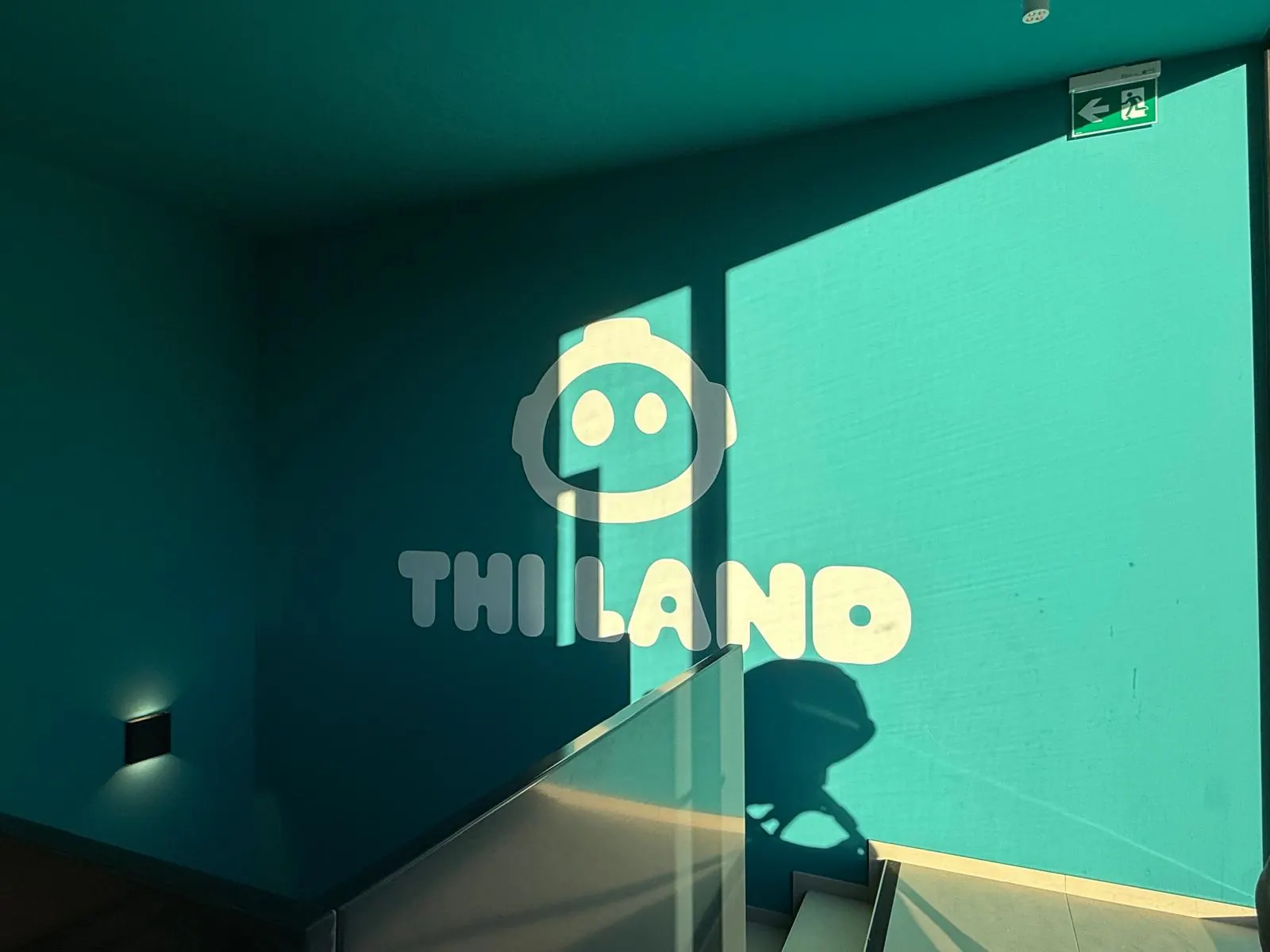 Il logo di THI LAND proiettato dal sole sulla parete turchese dell'ingresso